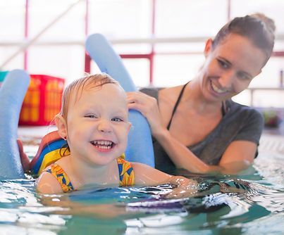 Child in pool with instructor, using flotation device, smiling. Water, indoor setting.
