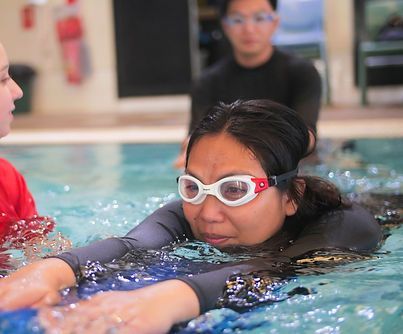 Woman in a pool wearing goggles, practicing swimming with instructors in the background.