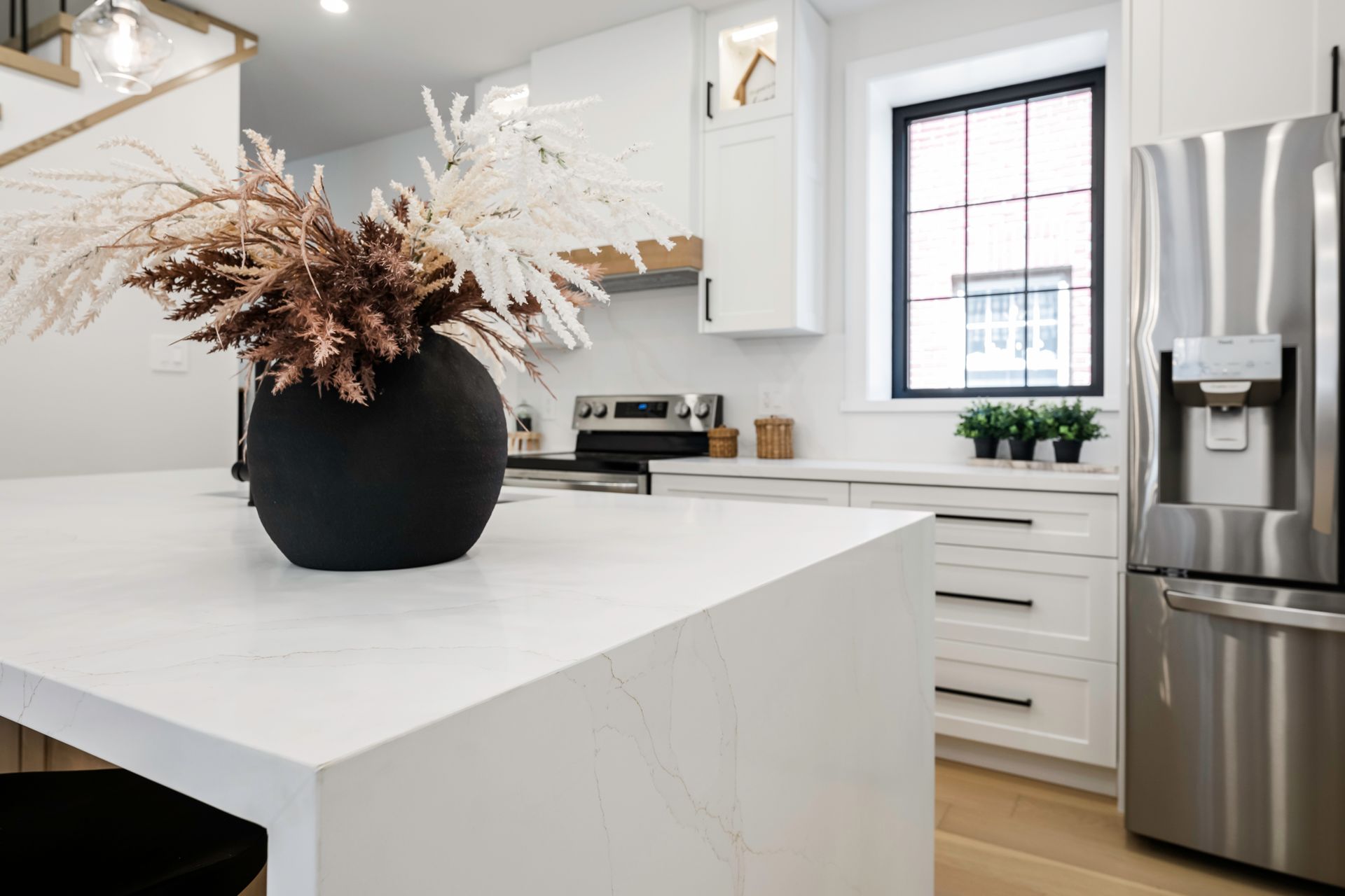 Modern white kitchen with island, black vase of dried flowers, stainless steel fridge, window.