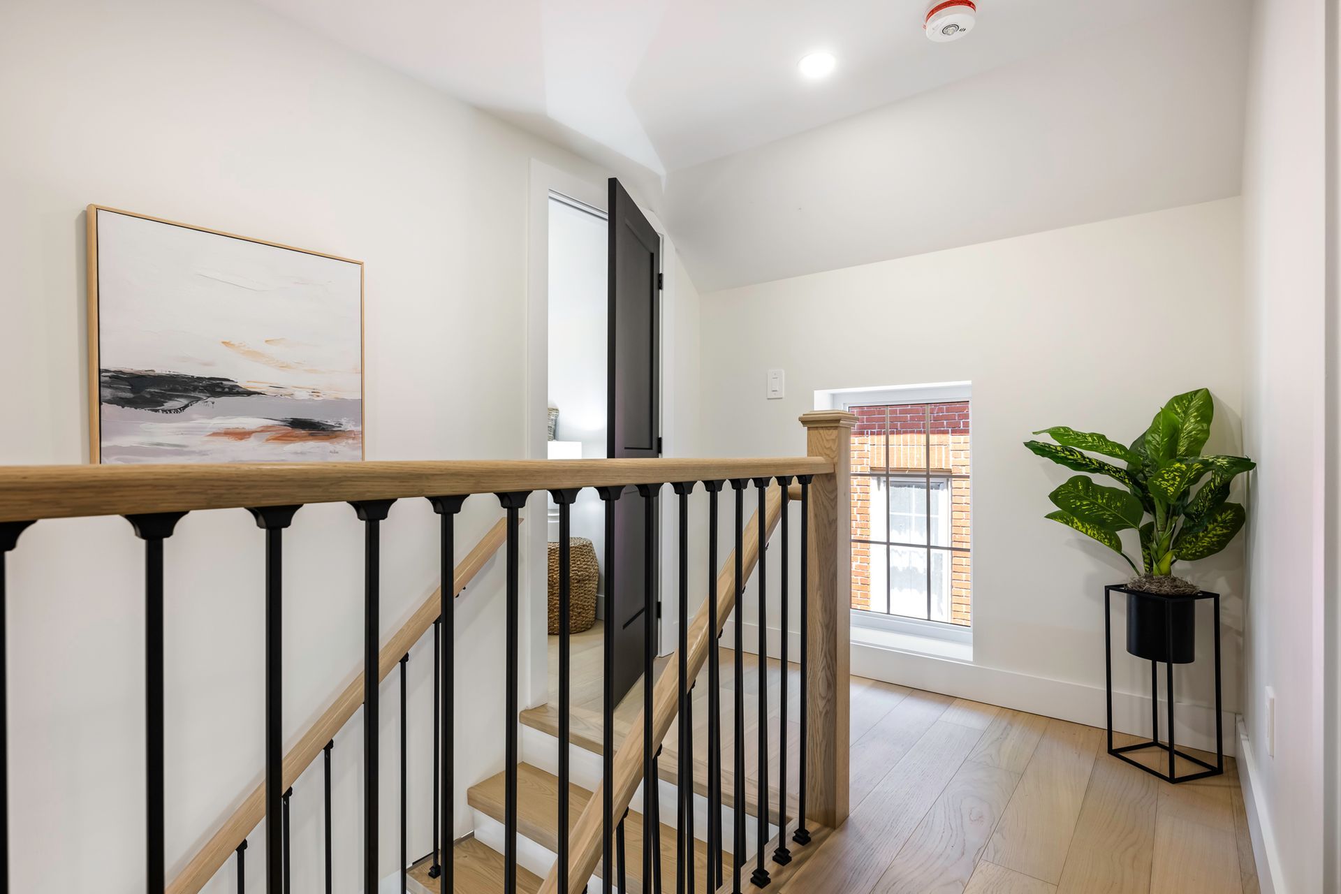 Hallway with stairs and black railing, light wood floors, artwork, potted plant, and a window.