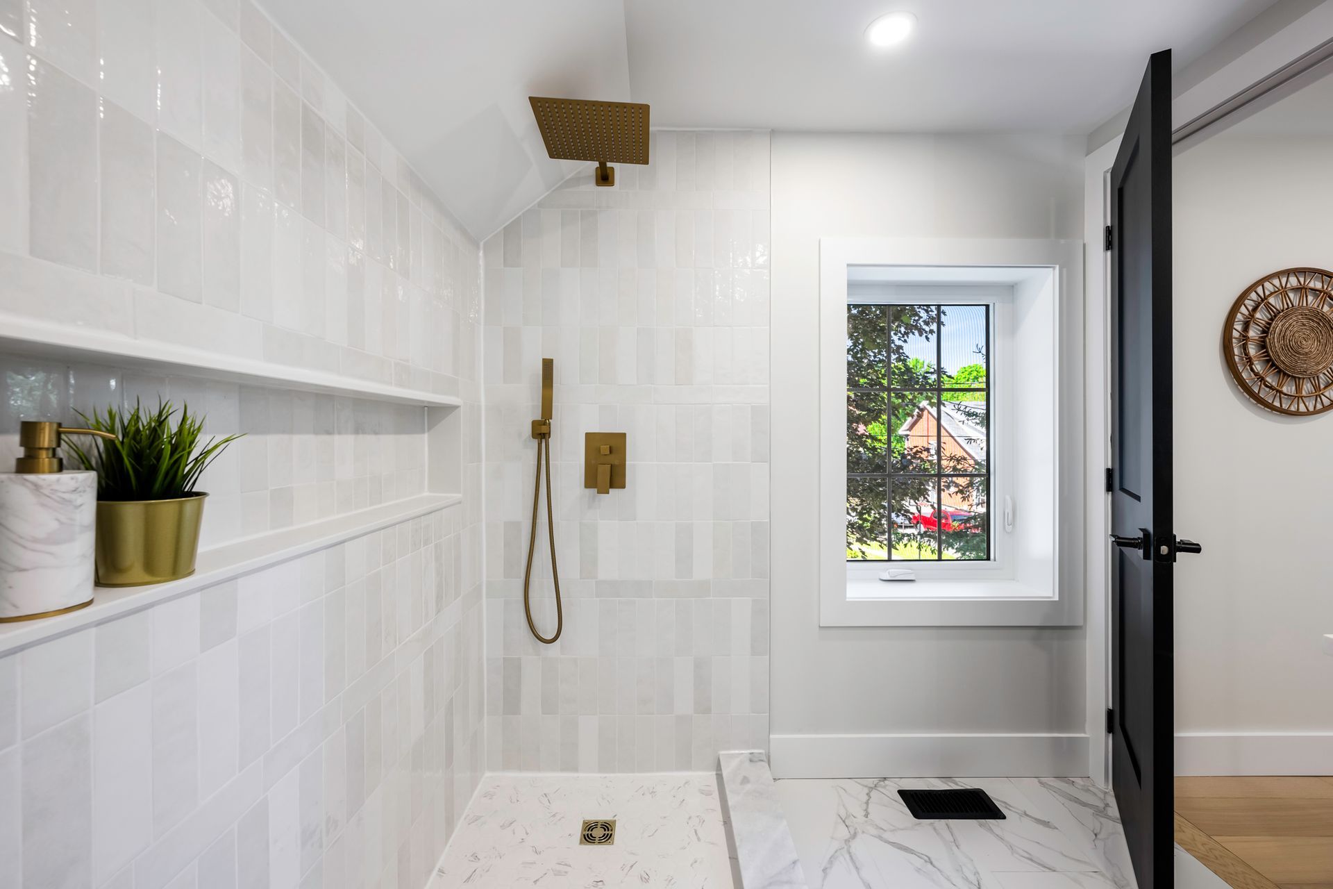 Modern white tiled shower with gold fixtures, a small window, and a dark door.