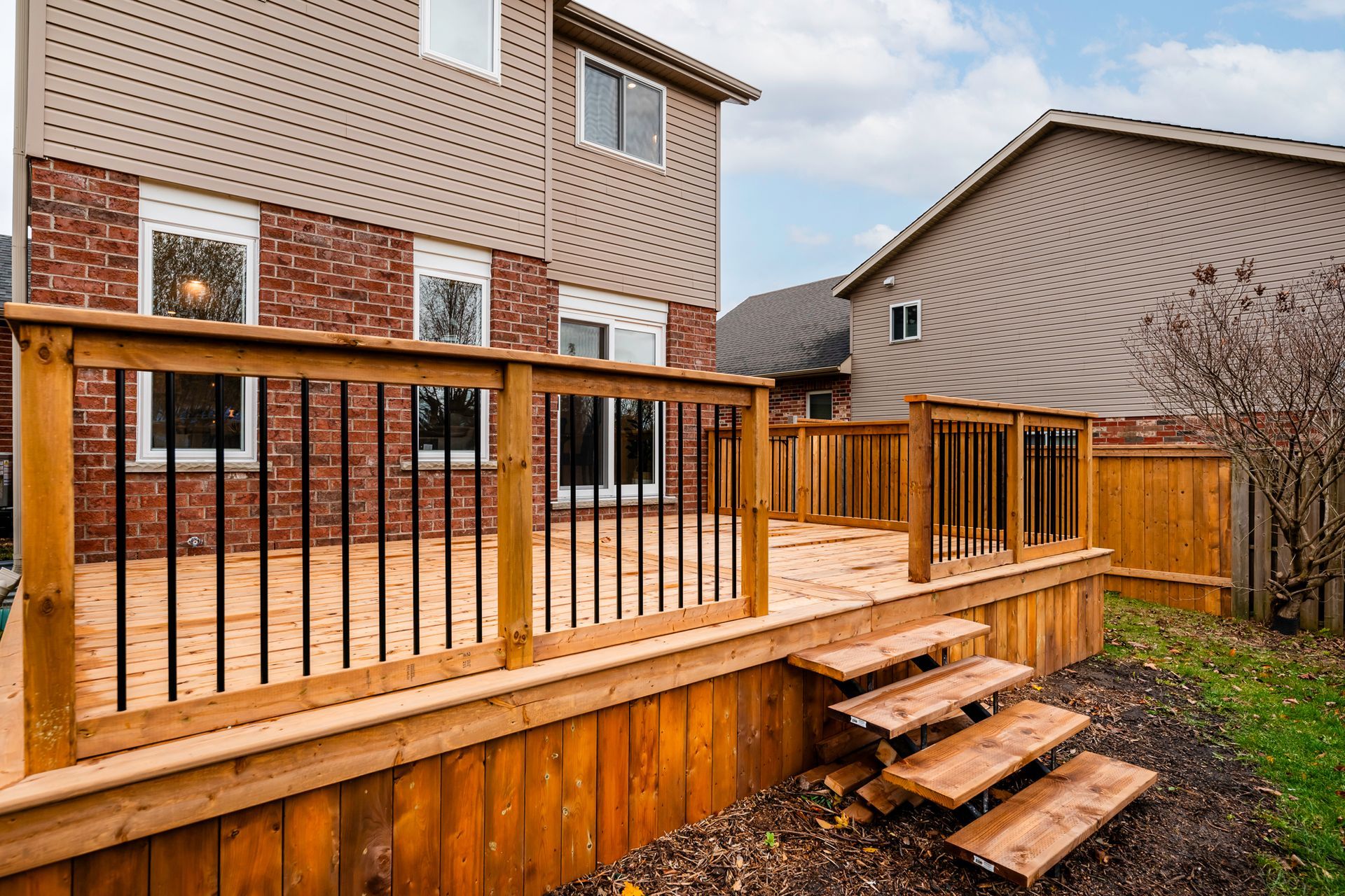 Wooden deck with black railings and steps leading down to a yard, against a brick and beige house.