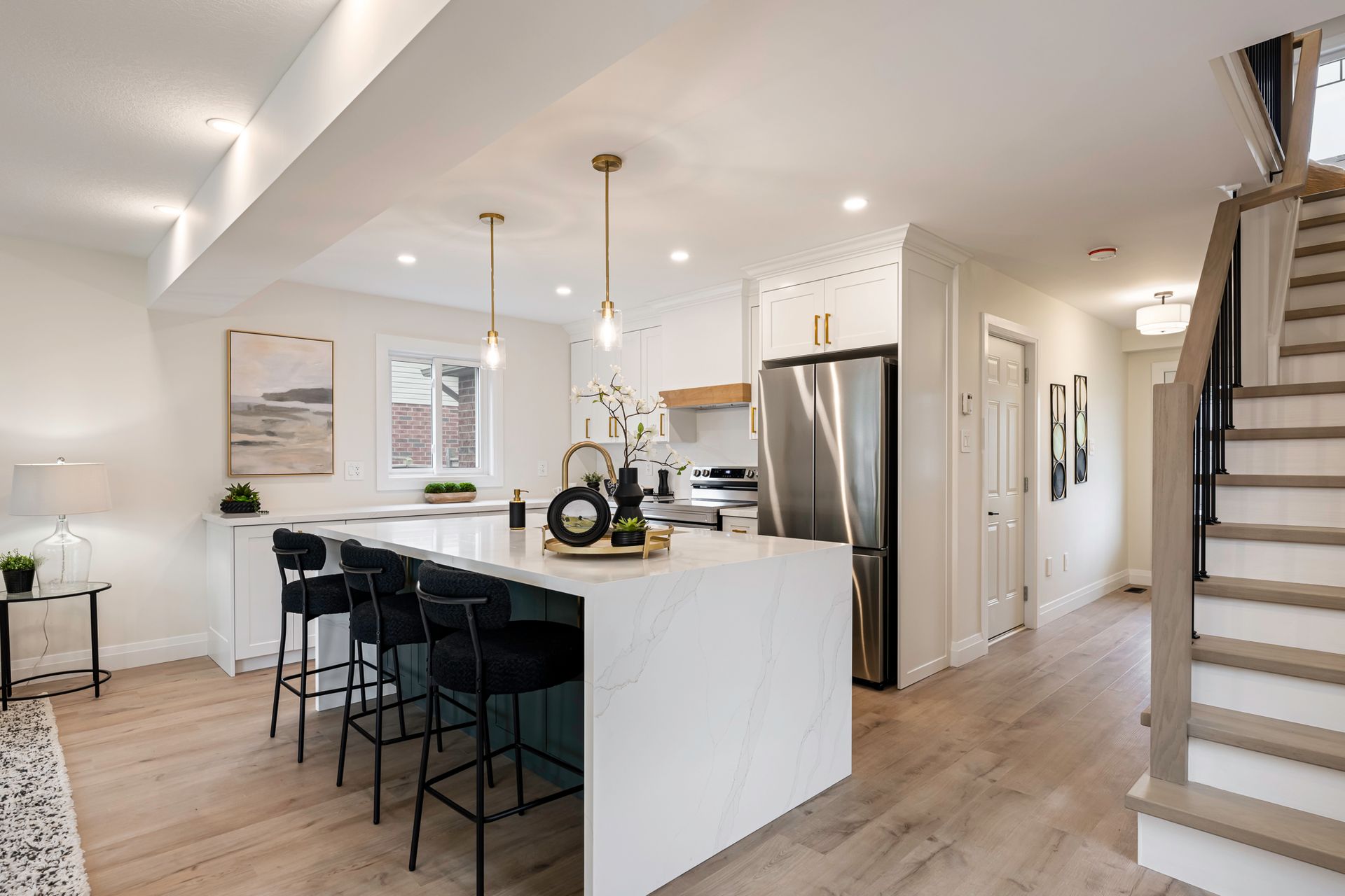 Modern kitchen with white countertops, island with black stools, stainless steel appliances, and wooden staircase.