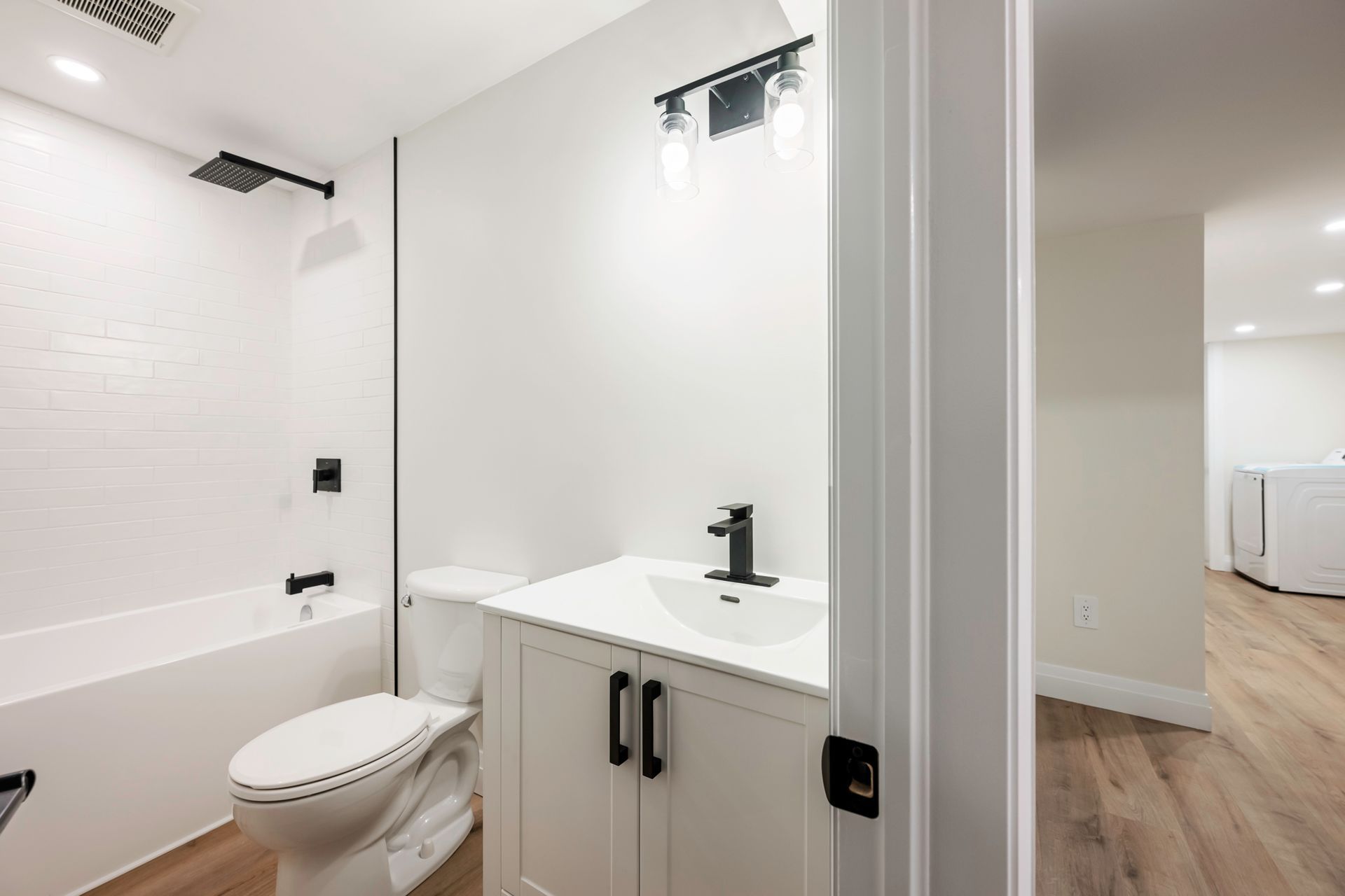 Bathroom with white walls, black fixtures, and a white vanity. Light wood-look flooring. Open doorway to adjacent room.