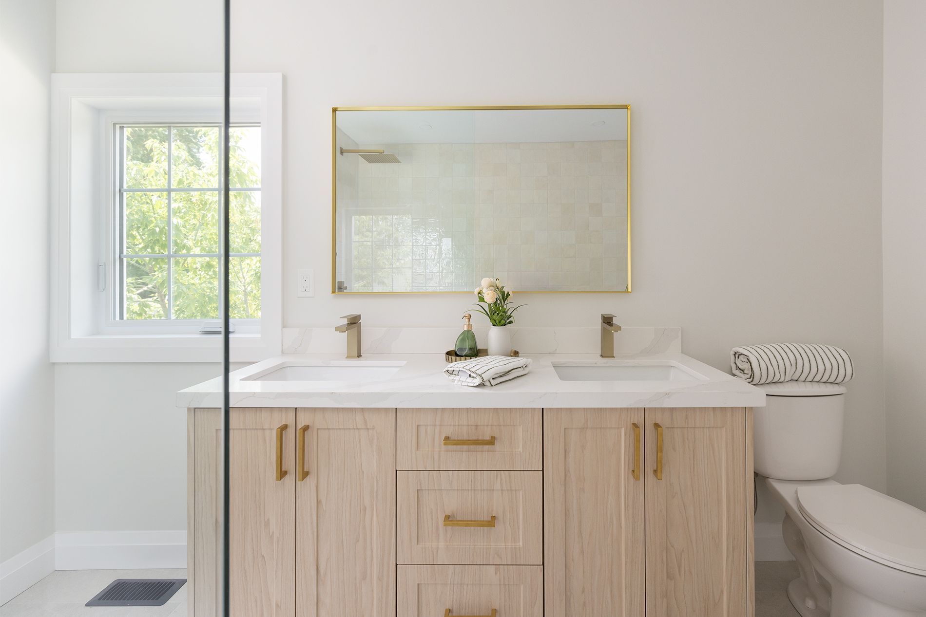Light wood bathroom vanity with gold hardware, marble countertop, and rectangular gold-framed mirror.