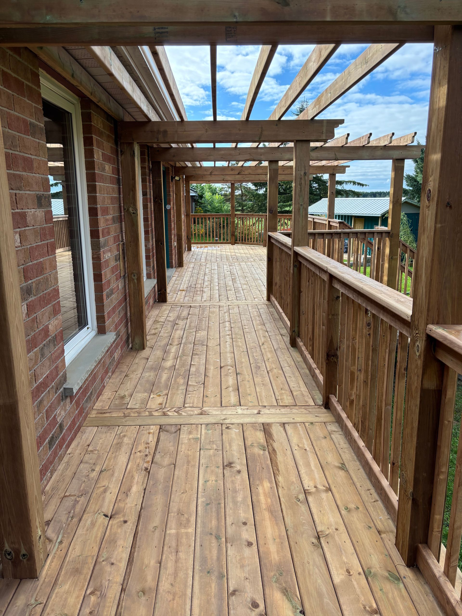 Wooden deck with pergola, brick wall on one side, railings, and a blue sky.