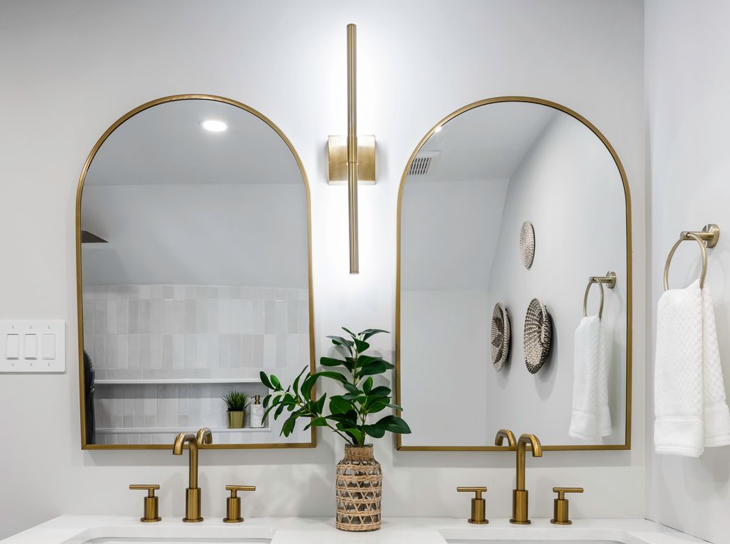 Bathroom vanity with two arched gold-framed mirrors, gold fixtures, and a decorative vase with greenery.