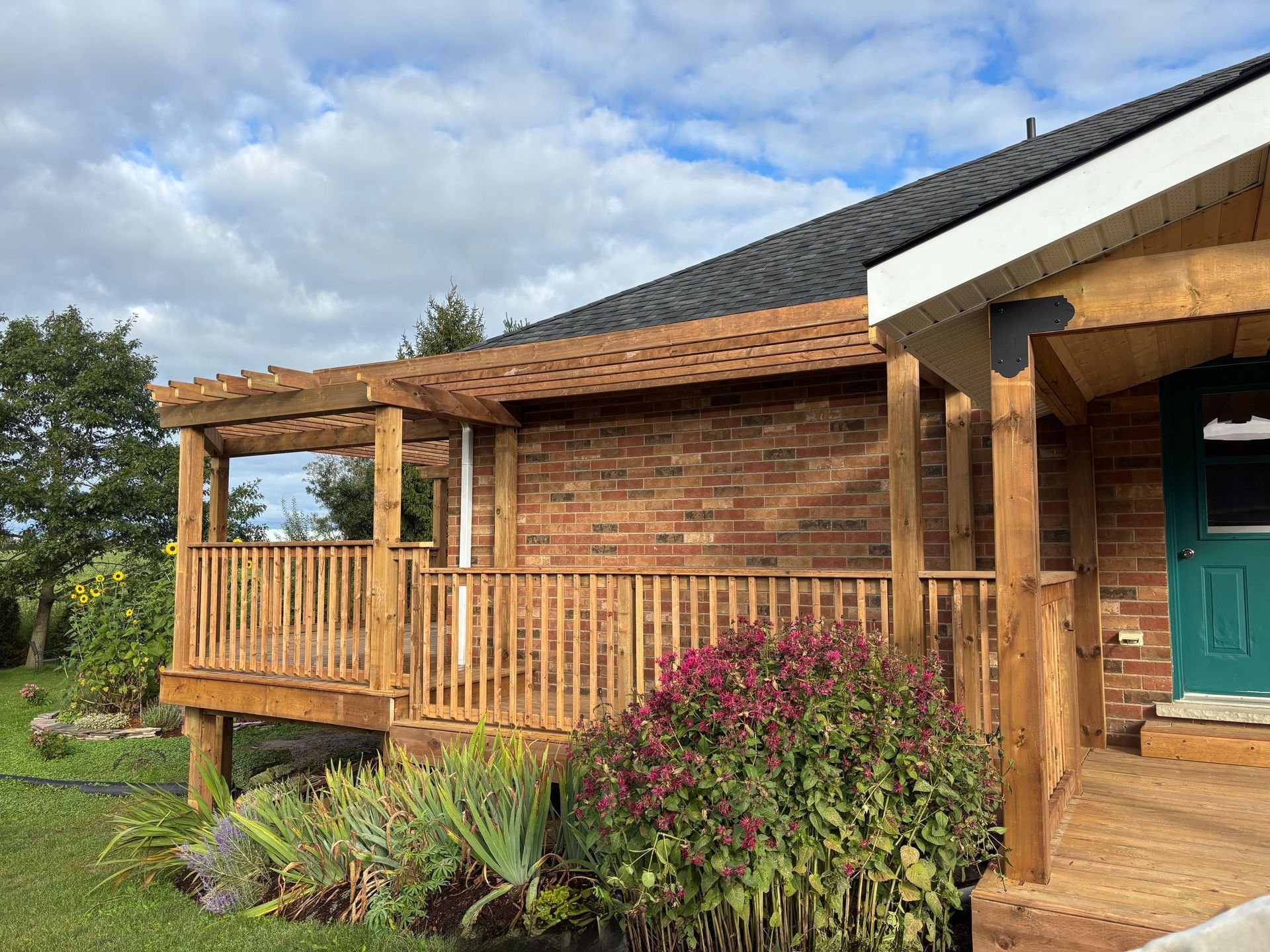 Wooden deck with pergola, brick wall, and green door under a cloudy sky.