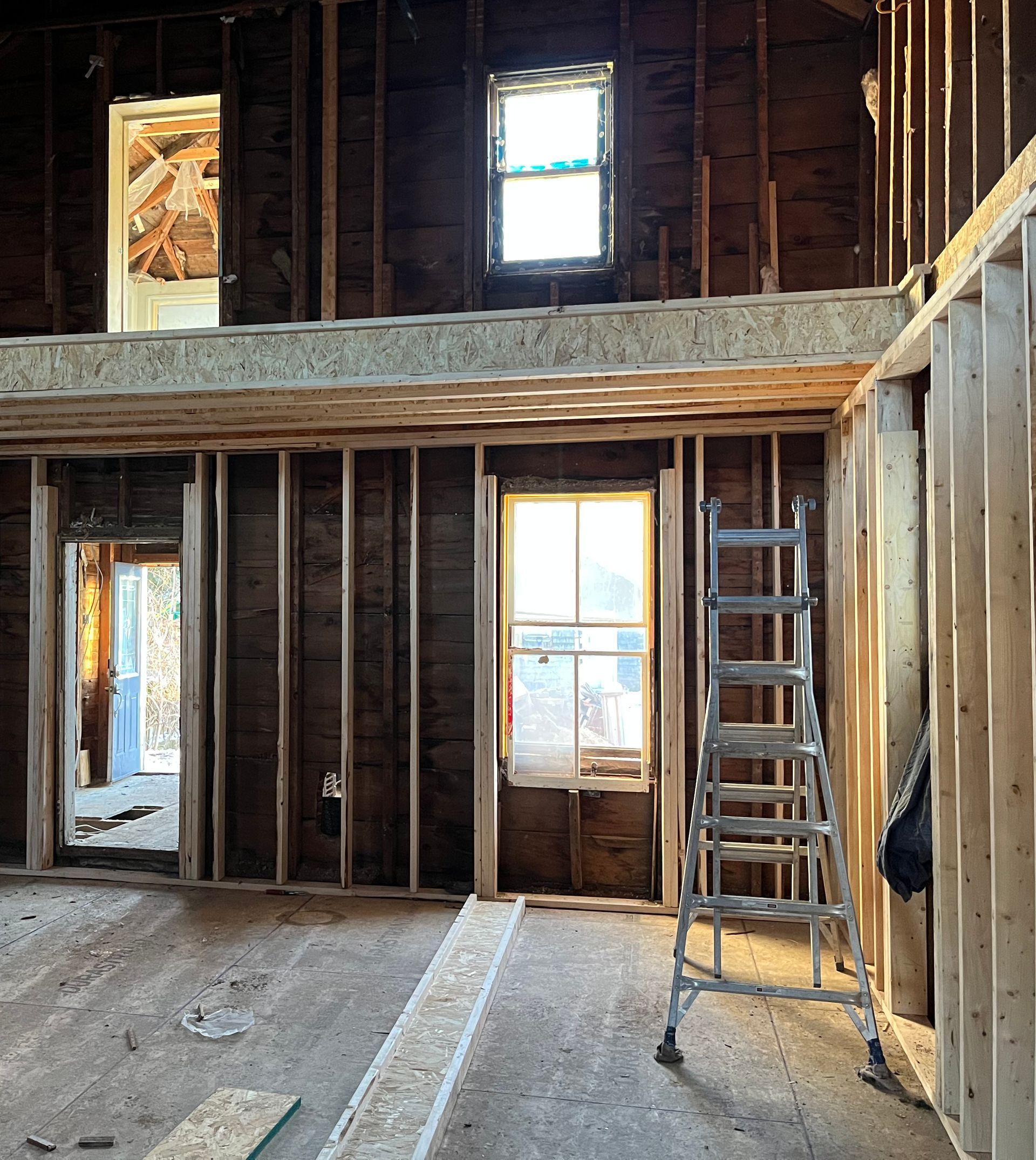 Interior of a house under renovation, exposed wooden studs, windows, and a ladder.
