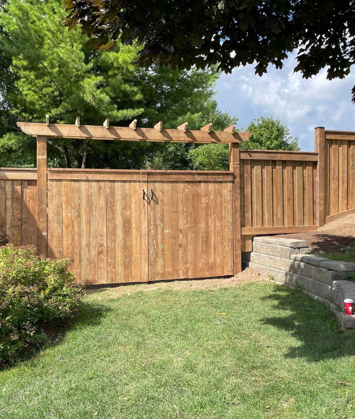 Wooden gate in a backyard with a pergola, flanked by a wooden fence and retaining wall, green grass and trees.