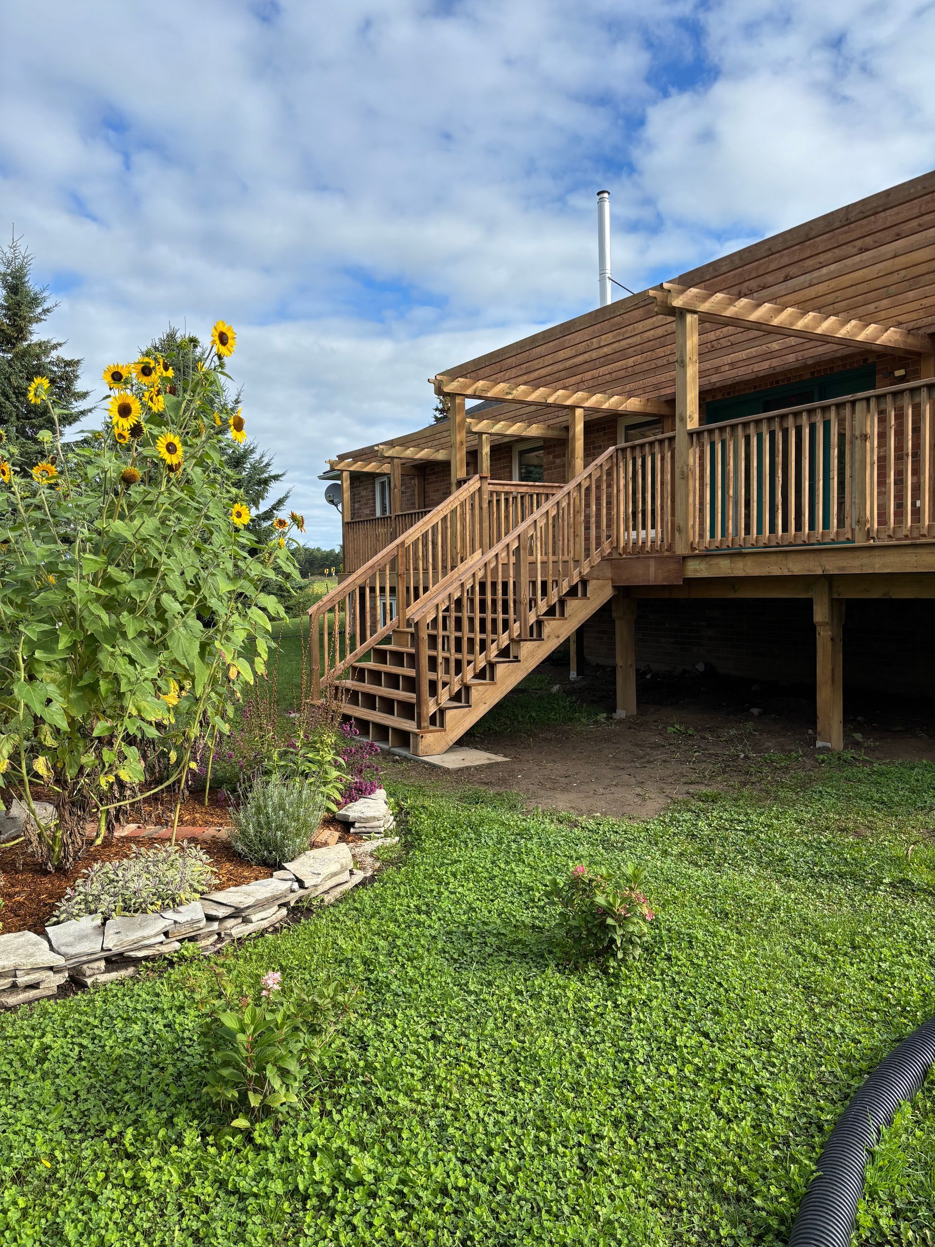 Wooden deck with stairs, pergola, and sunflowers on a grassy lawn under a partly cloudy sky.