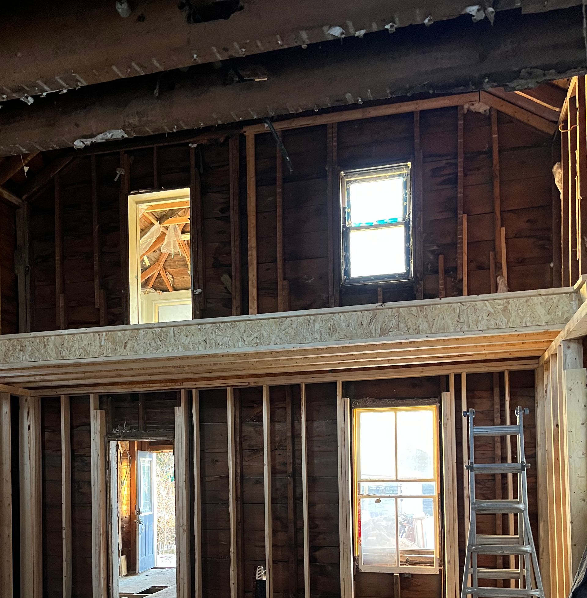 Interior view of a house under construction with exposed wooden framing and windows. A ladder leans against the wall.