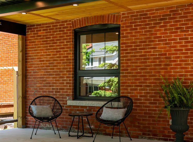 Brick wall with black-framed window, two black wire chairs, small table, and potted plant on a patio.