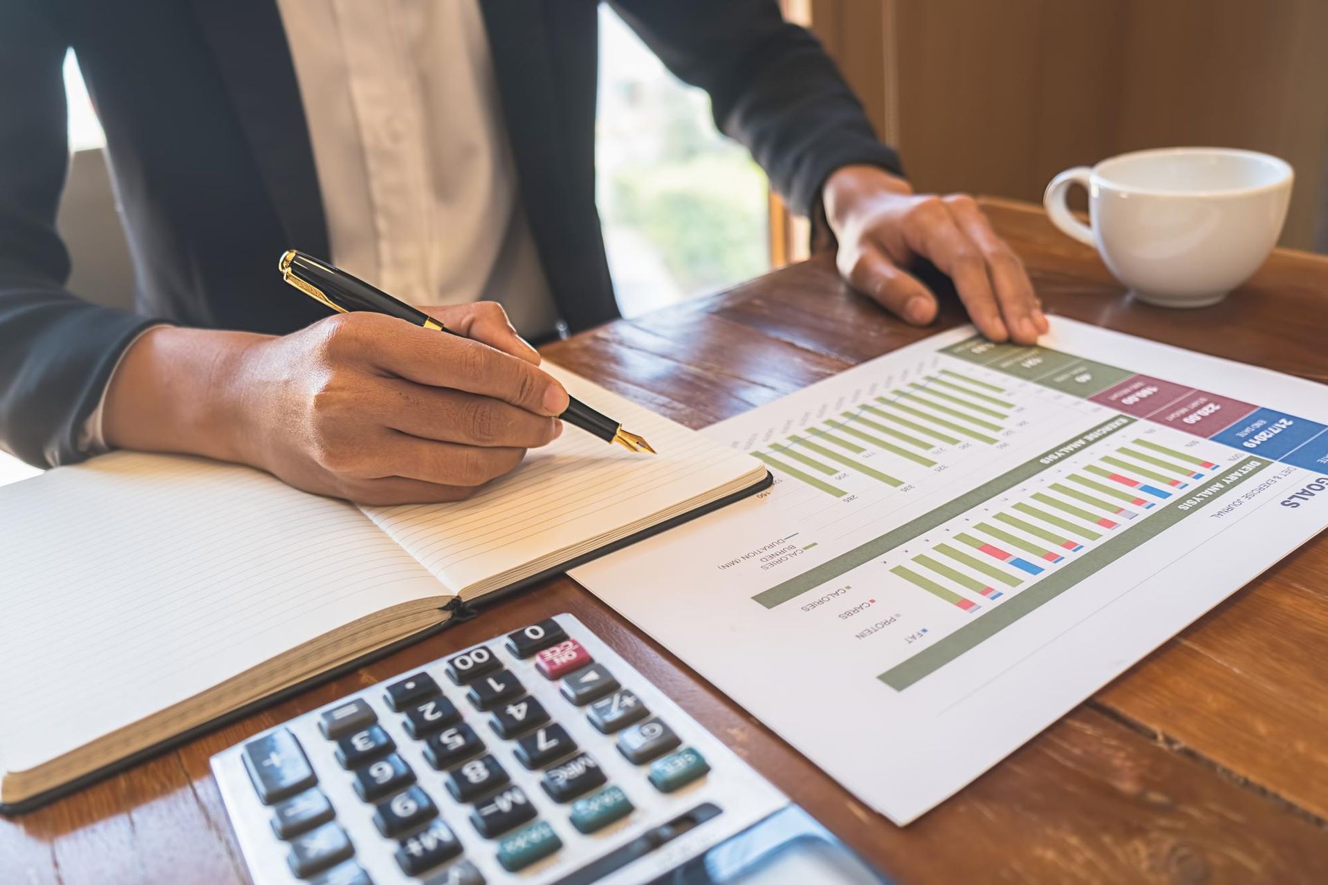 Person in blazer writing on a notepad, reviewing financial charts and using a calculator at a wooden table.