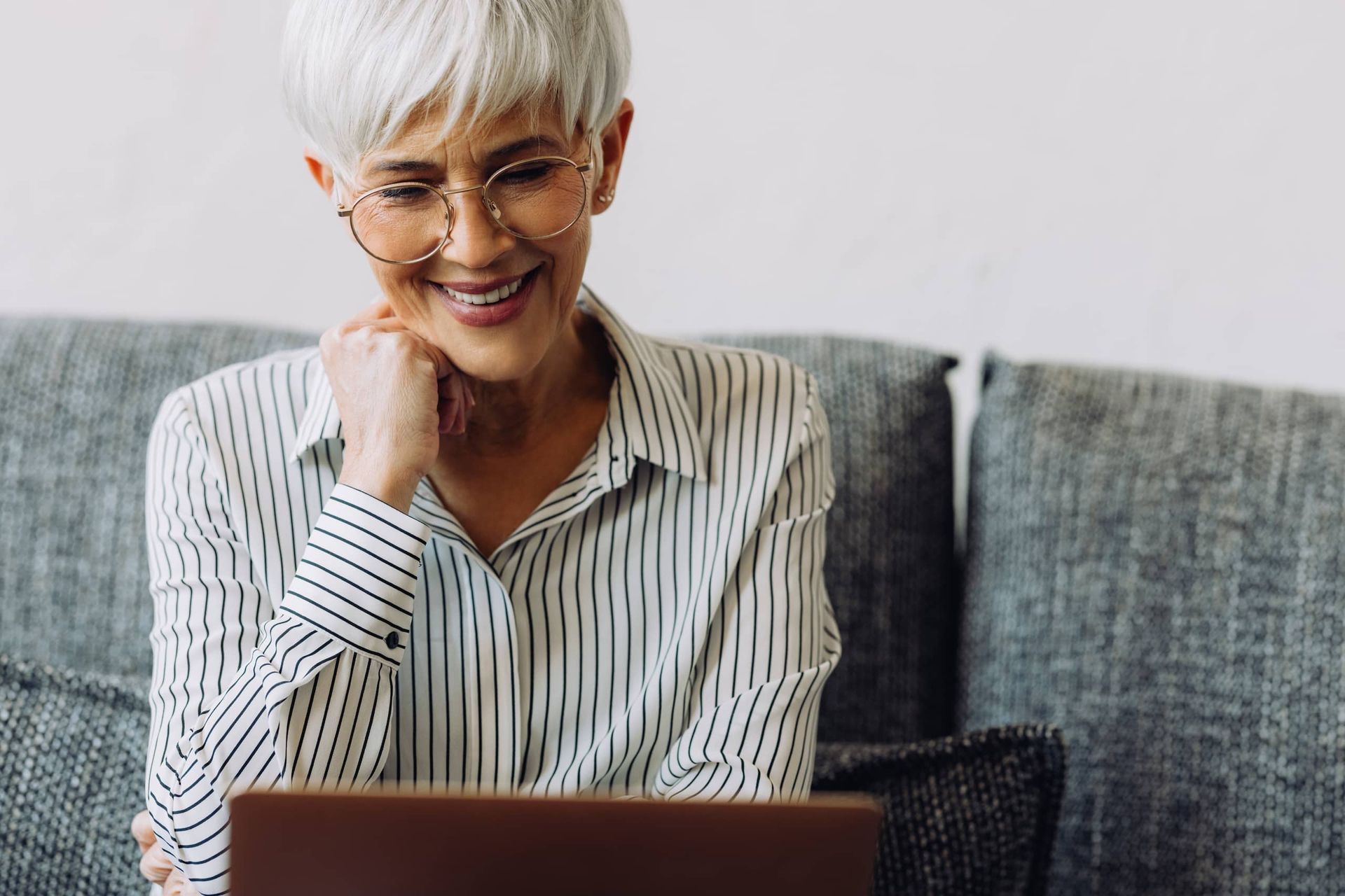 Woman with glasses smiles while using a laptop on a gray couch.