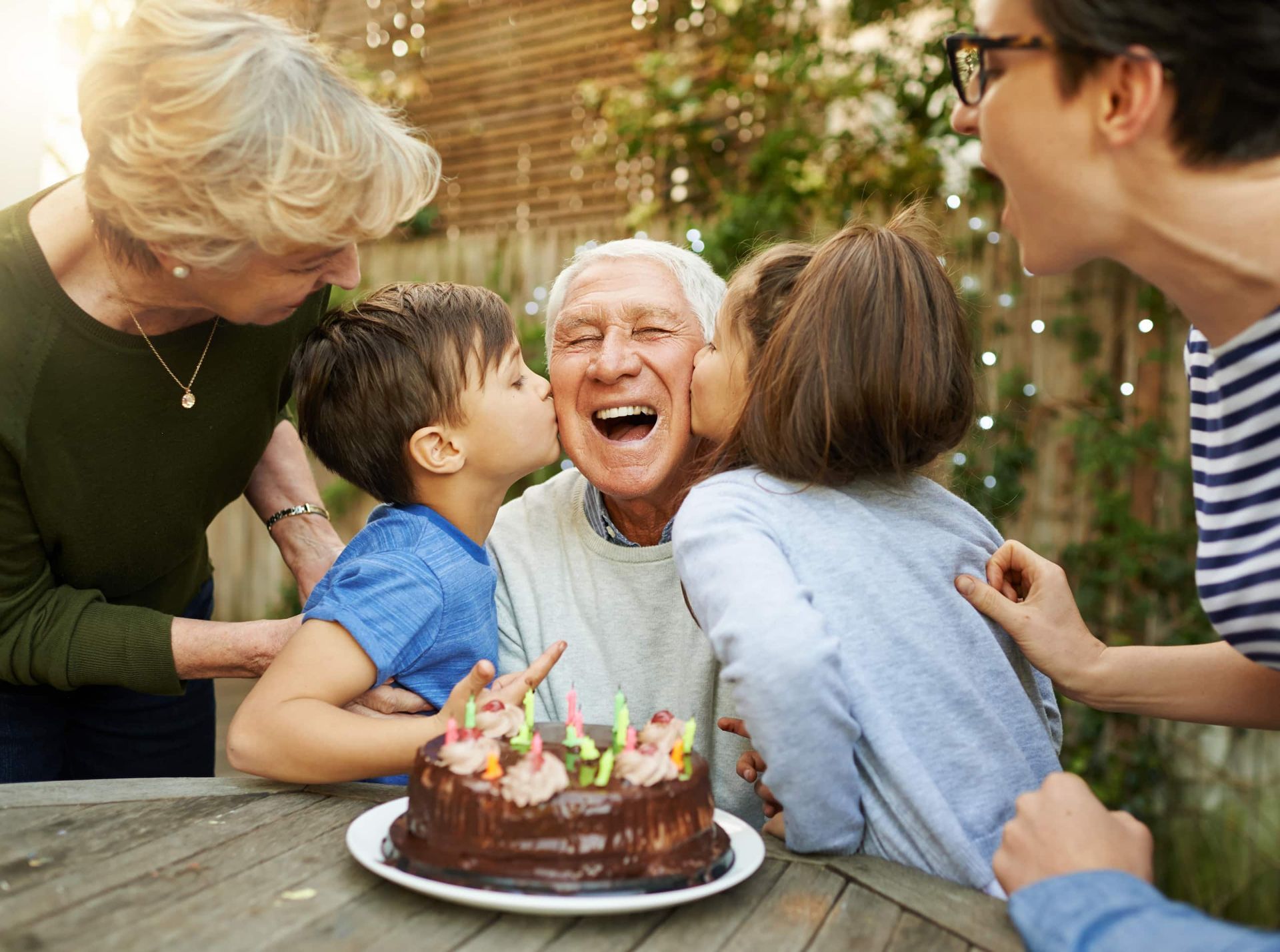 Family celebrating with a birthday cake outdoors; a man smiles as children and adults kiss his cheeks.