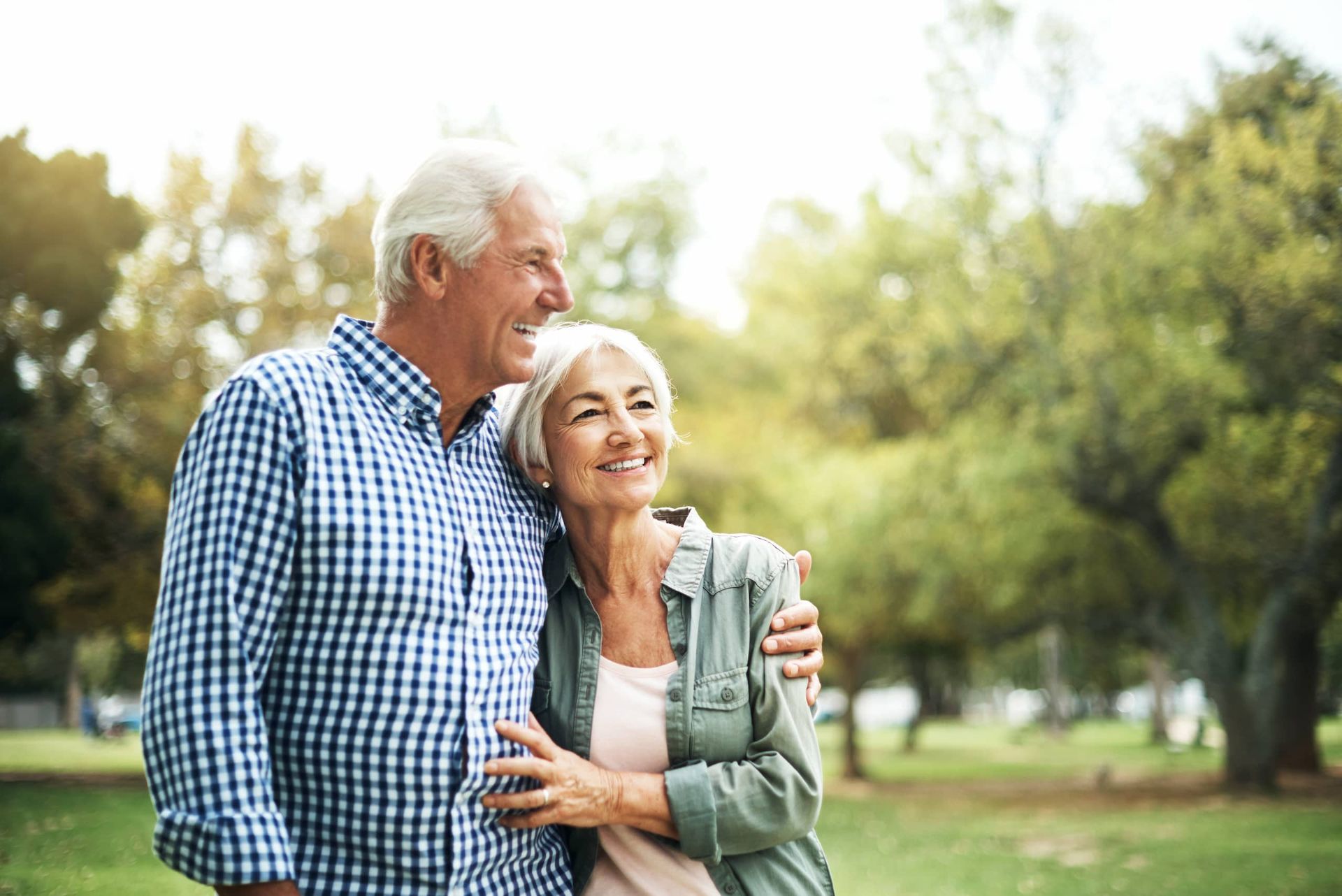 Elderly couple embraces, smiling, outdoors in a park. Man has arm around woman. Sunlight filters through trees.