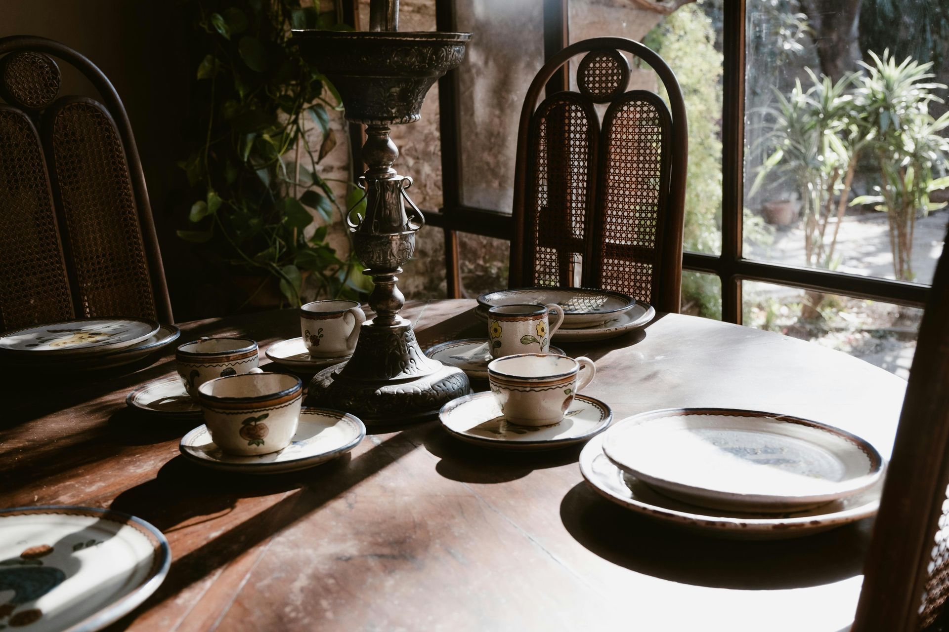 A wooden dining table set for a meal with teacups, plates, and a tall candle holder near a window.