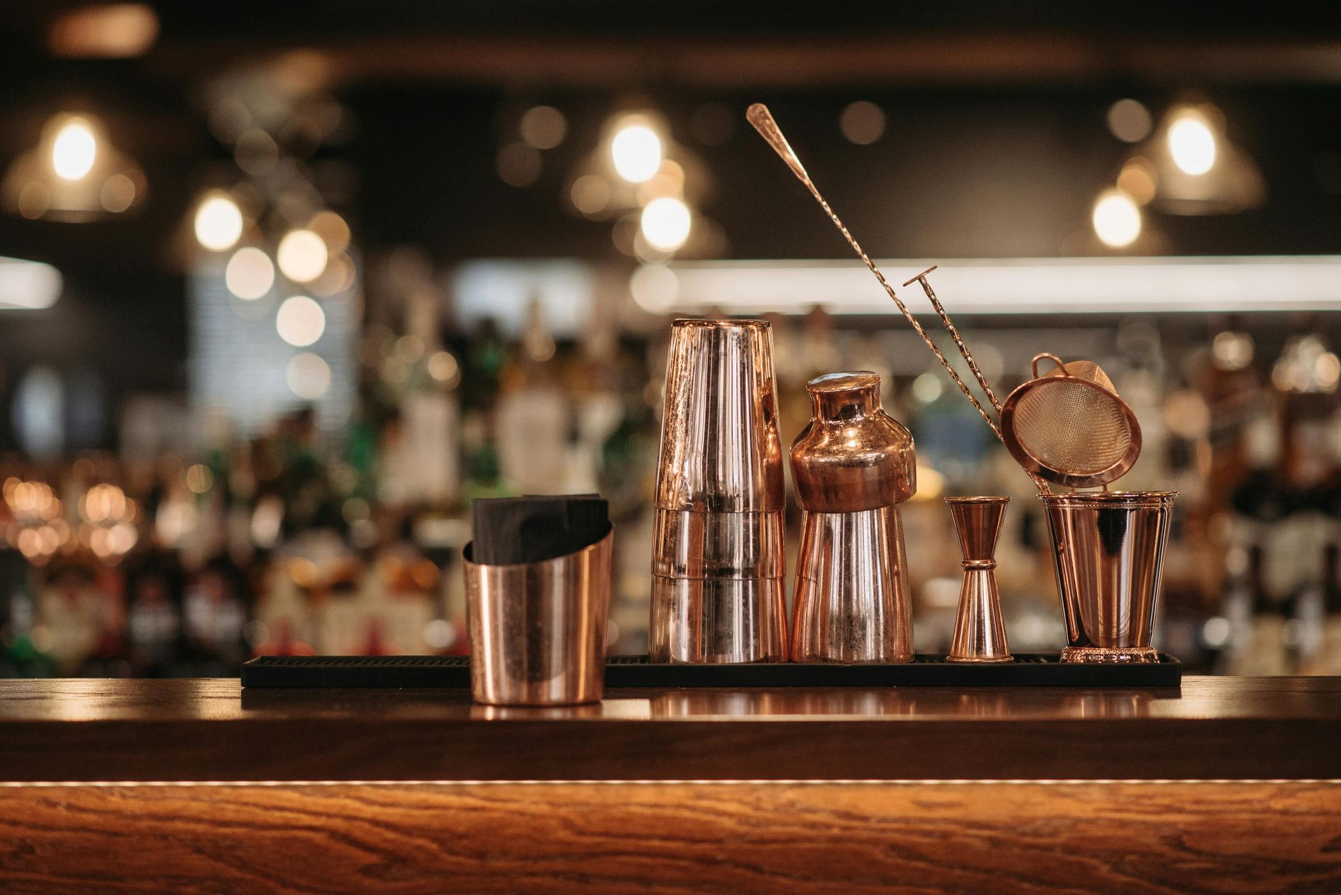 Copper bar tools on a wooden bar counter with a blurred background of liquor bottles and lights.