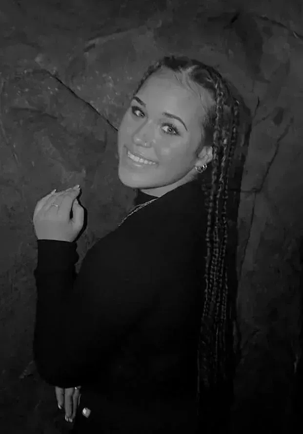 Woman with long braids smiles, leaning against a dark textured wall, wearing a black turtleneck.