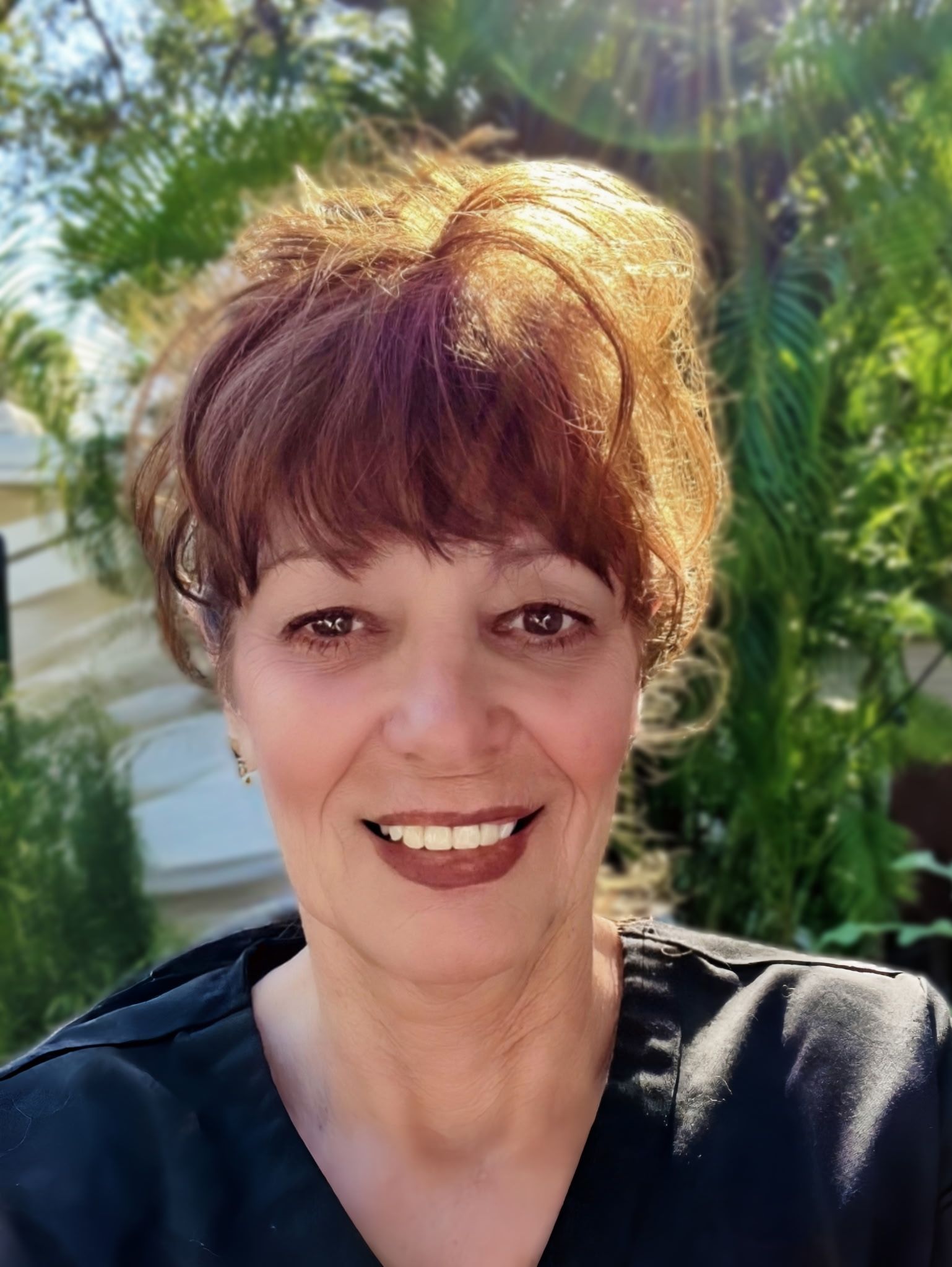 Woman with brown hair and bangs smiles at camera, wearing a white shirt and cross necklace.