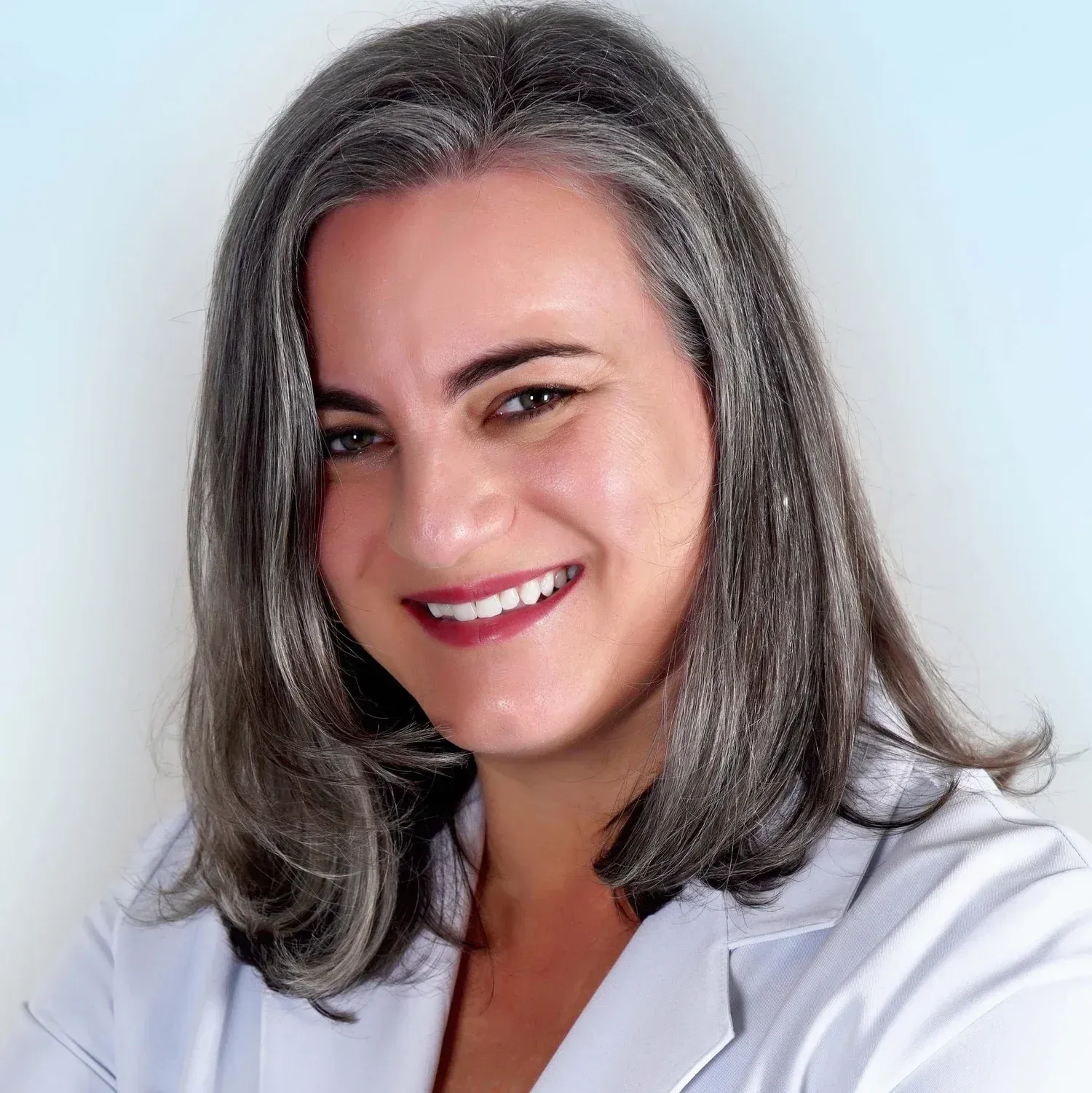 Woman with gray hair smiles in a white lab coat, light blue background.