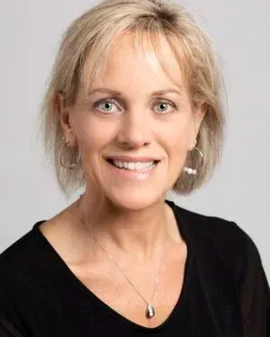 Woman with short blonde hair, smiling. Wearing a black top and a necklace. Studio setting.