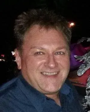 Man with short dark hair, smiling broadly, wearing a blue shirt, outdoors at night.