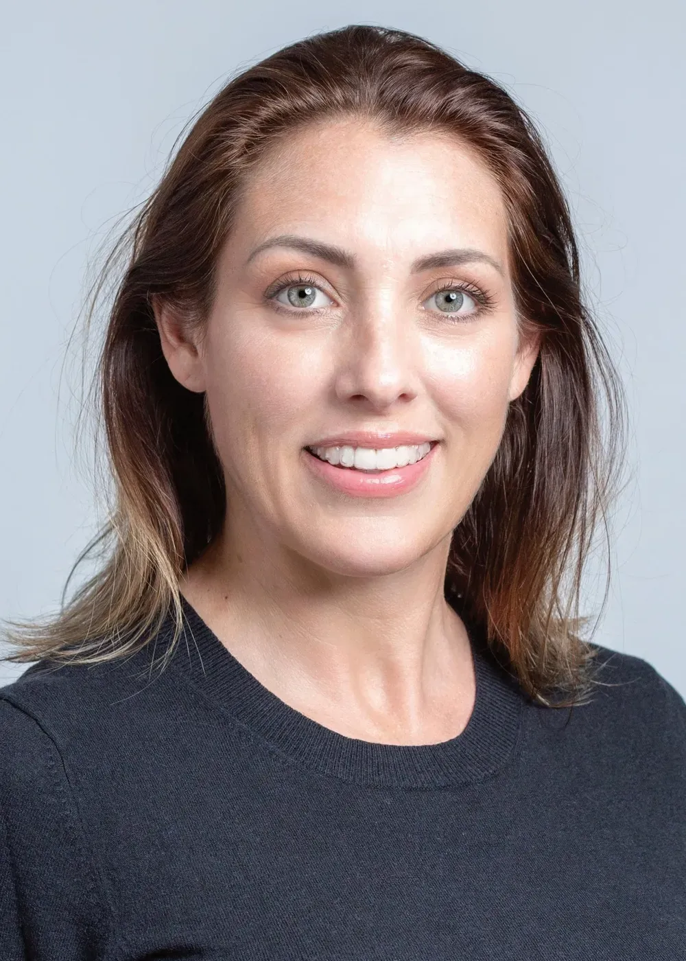 Woman with brown hair smiles, wearing a black shirt, against a gray background.