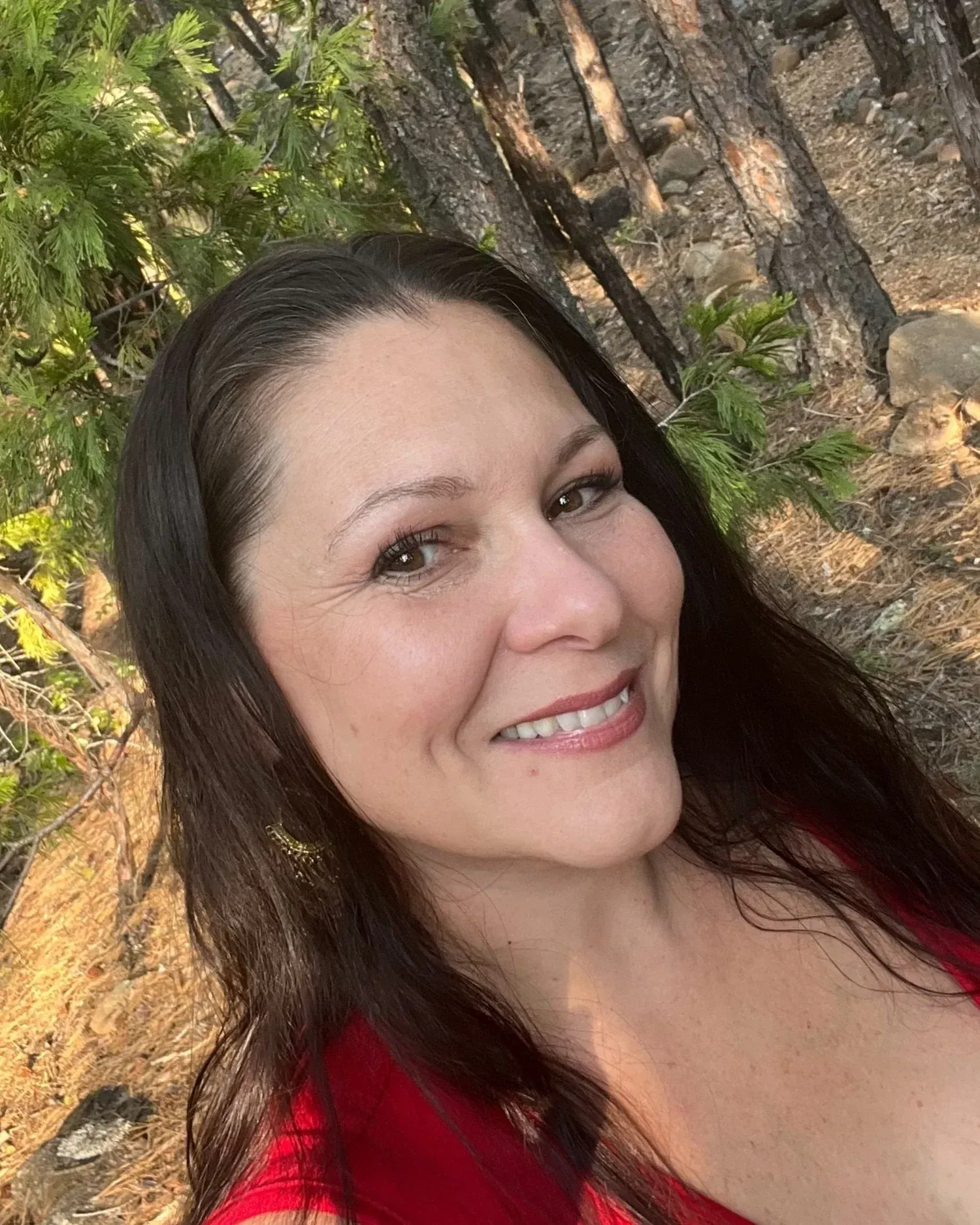 Woman with long dark hair smiles outdoors, wearing a red top. Forest background with trees.