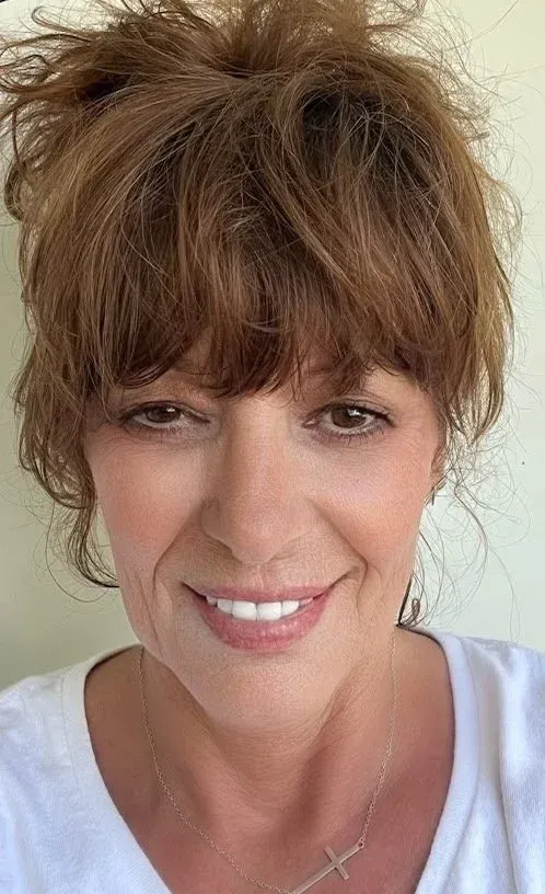 Woman with brown hair and bangs smiles at camera, wearing a white shirt and cross necklace.