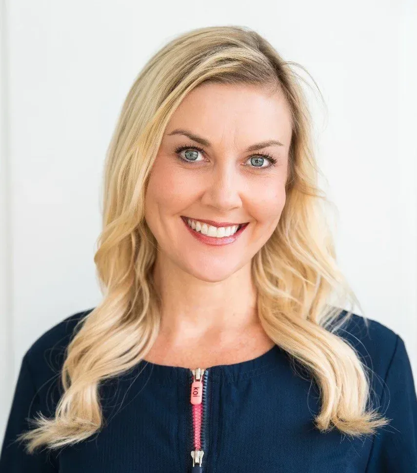 Blonde woman with blue eyes smiles at the camera, wearing a navy blue top with a pink zipper against a white background.