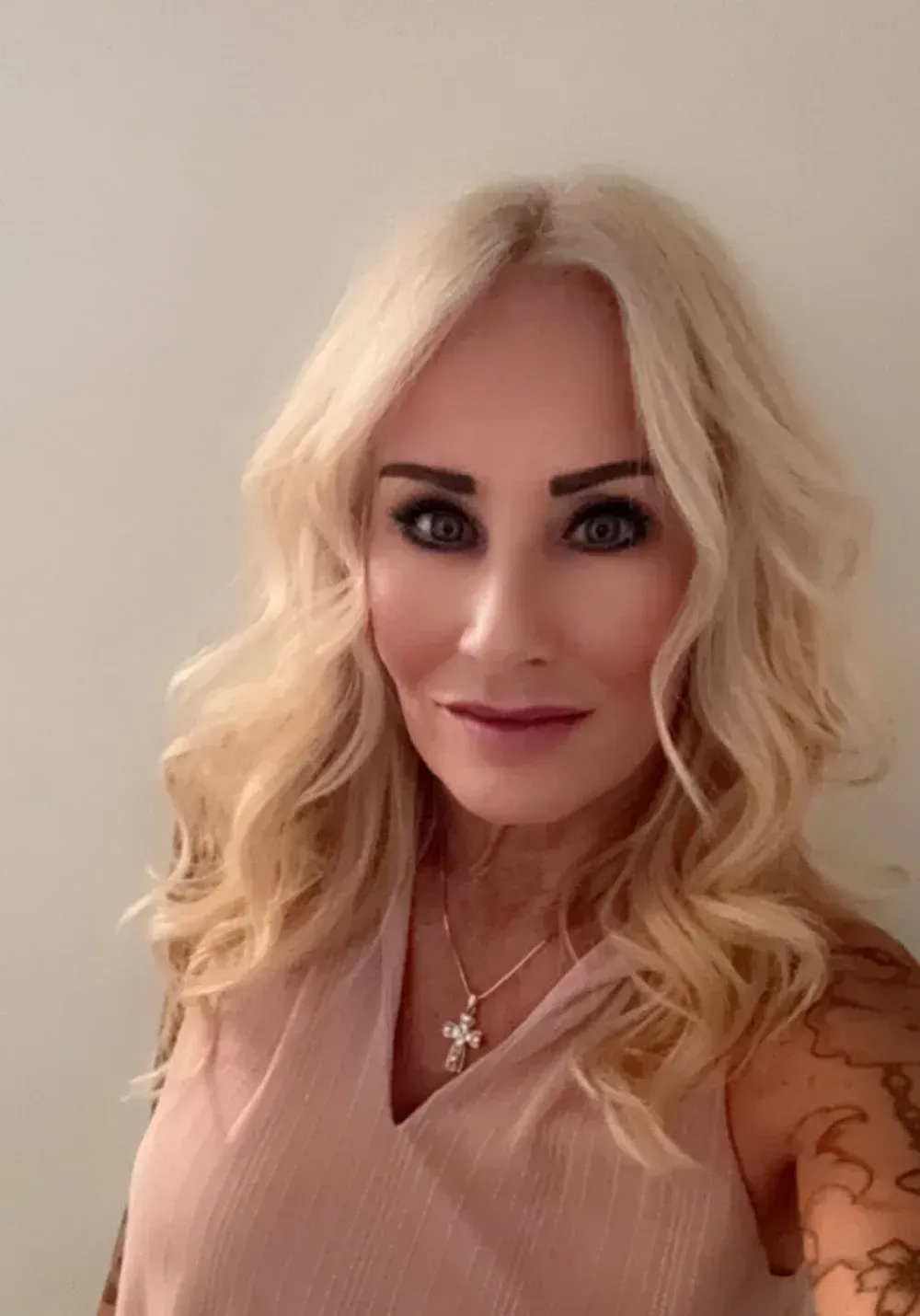 Blonde woman with wavy hair, wearing a pink top and cross necklace, smiles at the camera.