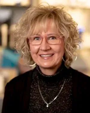 Woman with blonde curly hair and pink glasses smiles, wearing black turtleneck and blazer.