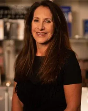 Woman with long brown hair smiling at the camera, wearing a black top, in a store setting.