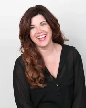 Woman with long brown hair laughs, wearing a black shirt, against a light background.