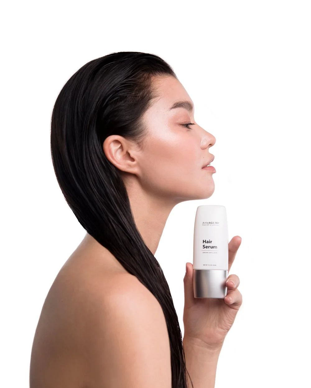 Woman holds skincare product, looking up. Dark hair, light skin, white background.
