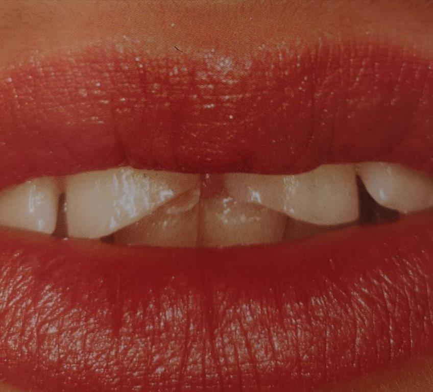 Primo piano di labbra rosse, con i denti che mordono leggermente il labbro. È visibile un piccolo dente anteriore scheggiato.