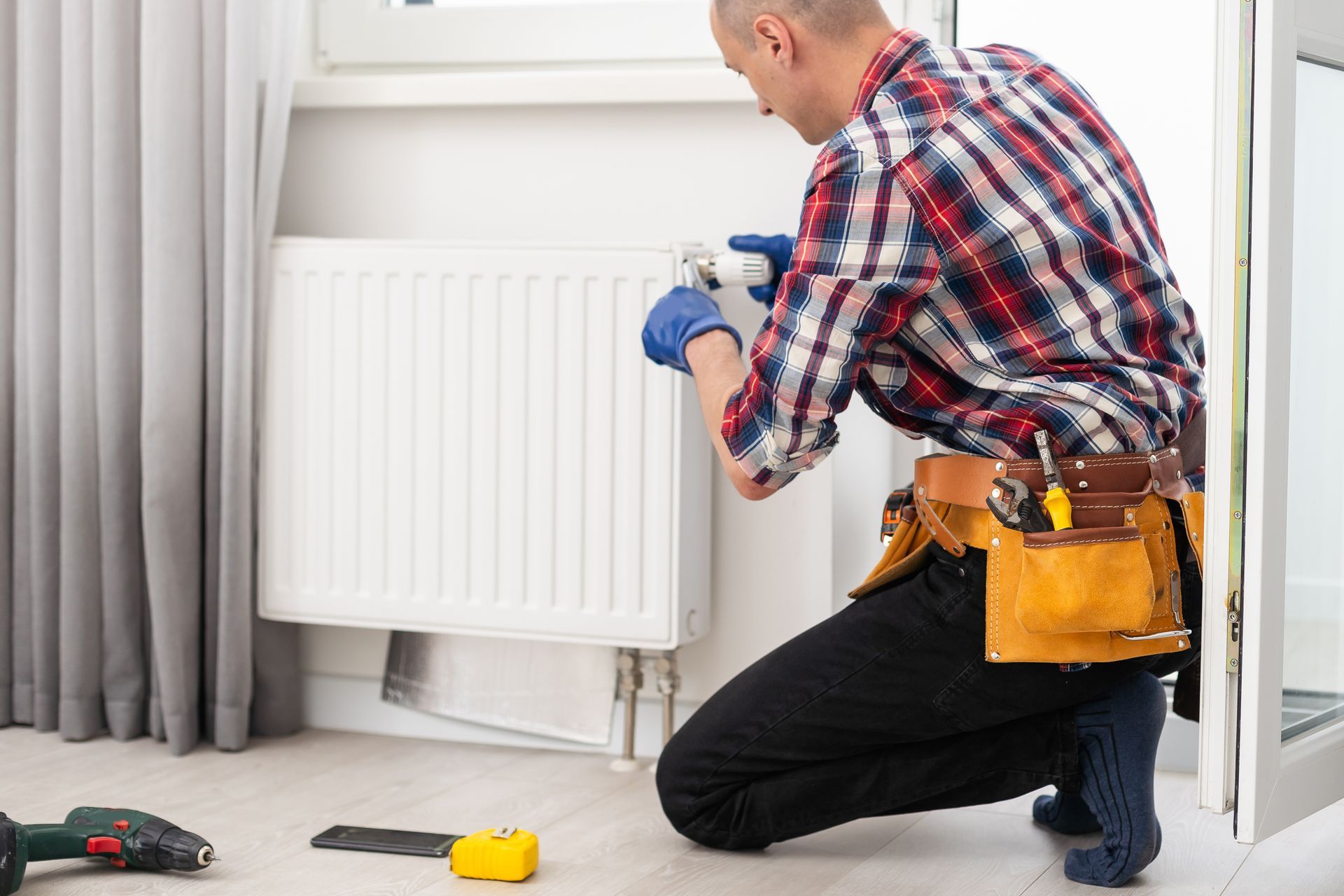 Plumber in plaid shirt kneeling, working on a white radiator near a window, with a tool belt. Plumber in plaid shirt kneeling, working on a white radiator near a window, with a tool belt.