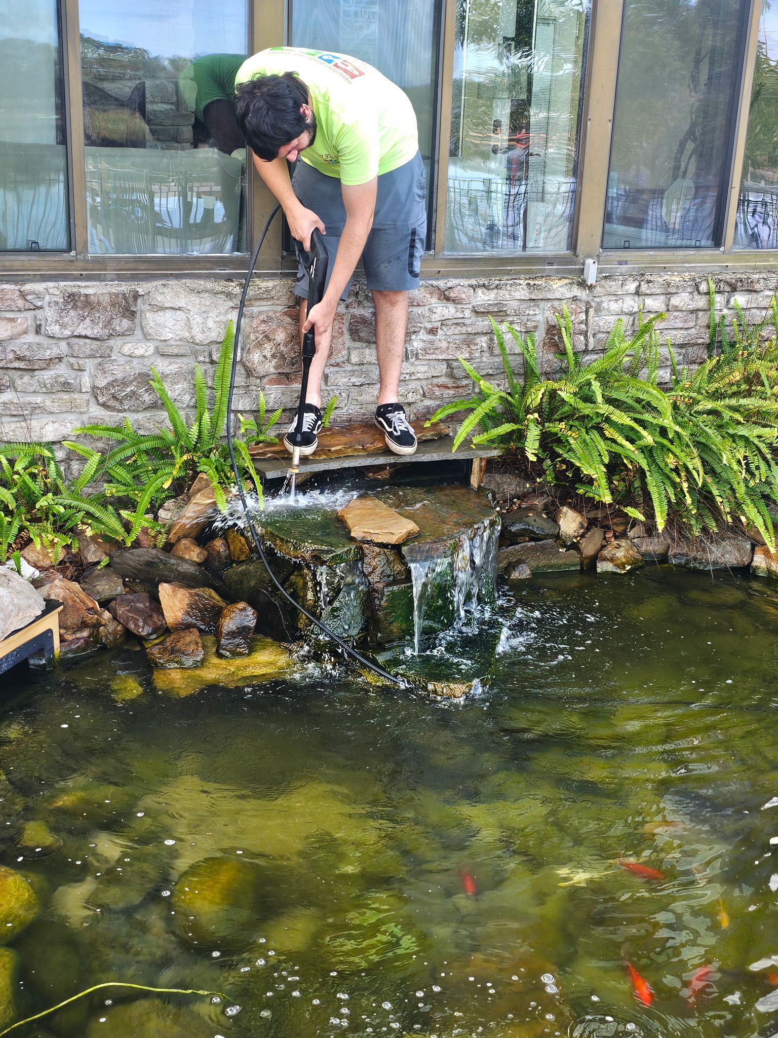Person leaning over a pond, pouring water from a bottle into a small waterfall beside plants and rocks.