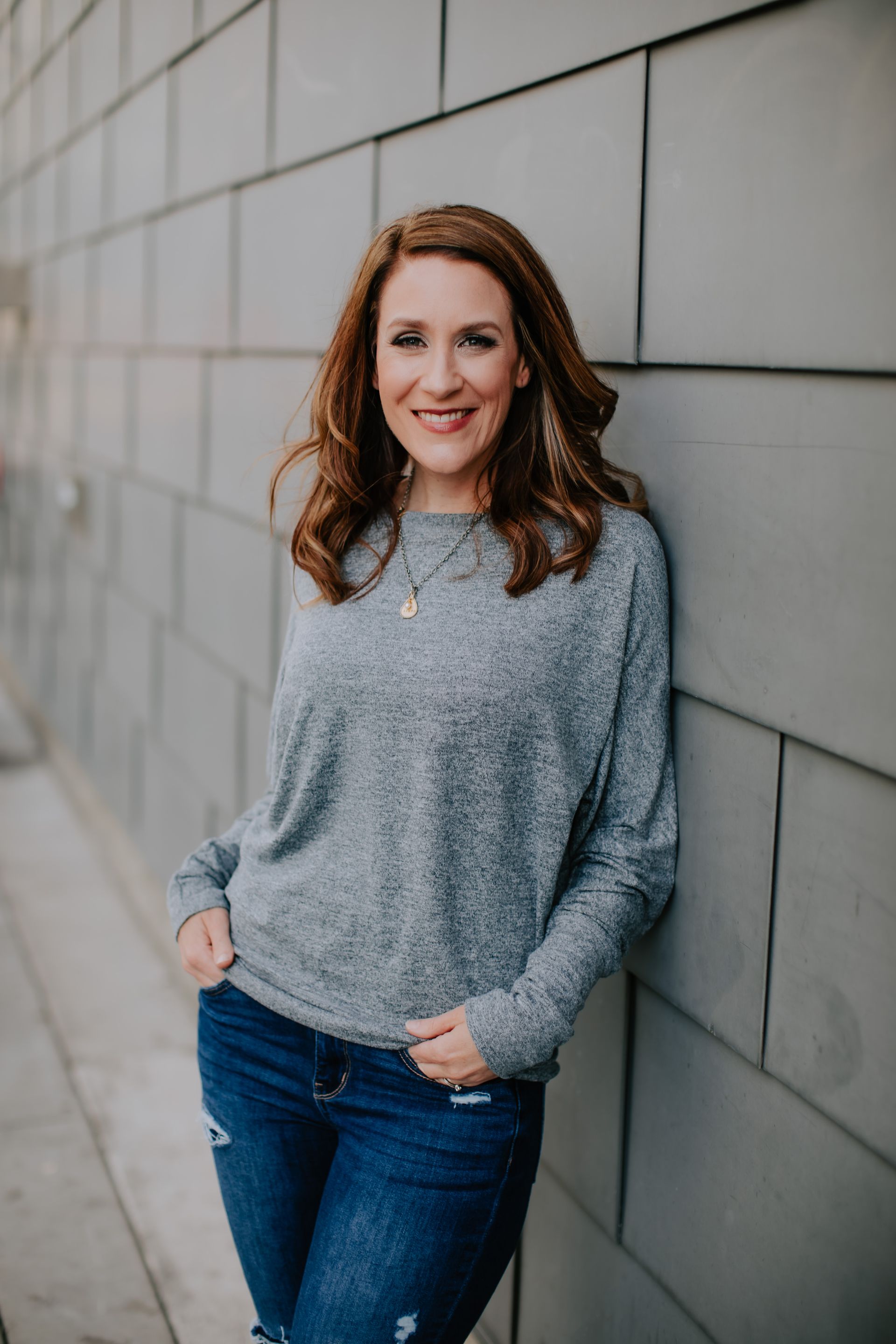 A woman in a grey sweater and blue jeans is leaning against a brick wall.