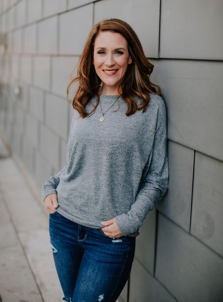 A woman in a grey sweater and blue jeans is leaning against a wall.