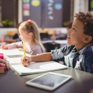 A young boy is sitting at a table in a classroom writing in a notebook.