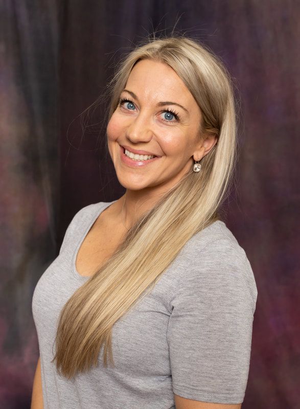 A woman with long blonde hair is wearing a grey shirt and smiling.