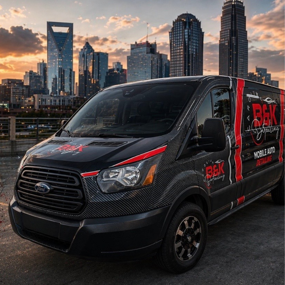 A person in a B&K Detailing uniform wipes the hood of a black BMW parked in front of a house.