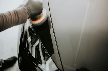 A person wearing a glove uses a power polisher on the headlight of a white car.