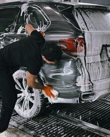 A person wearing a black shirt uses an orange sponge to wash a dark car covered in white soap suds at a car wash.