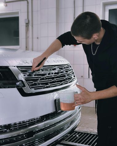 A person in a black outfit uses a detailing brush to clean the soapy front grille of a white SUV in a workshop.