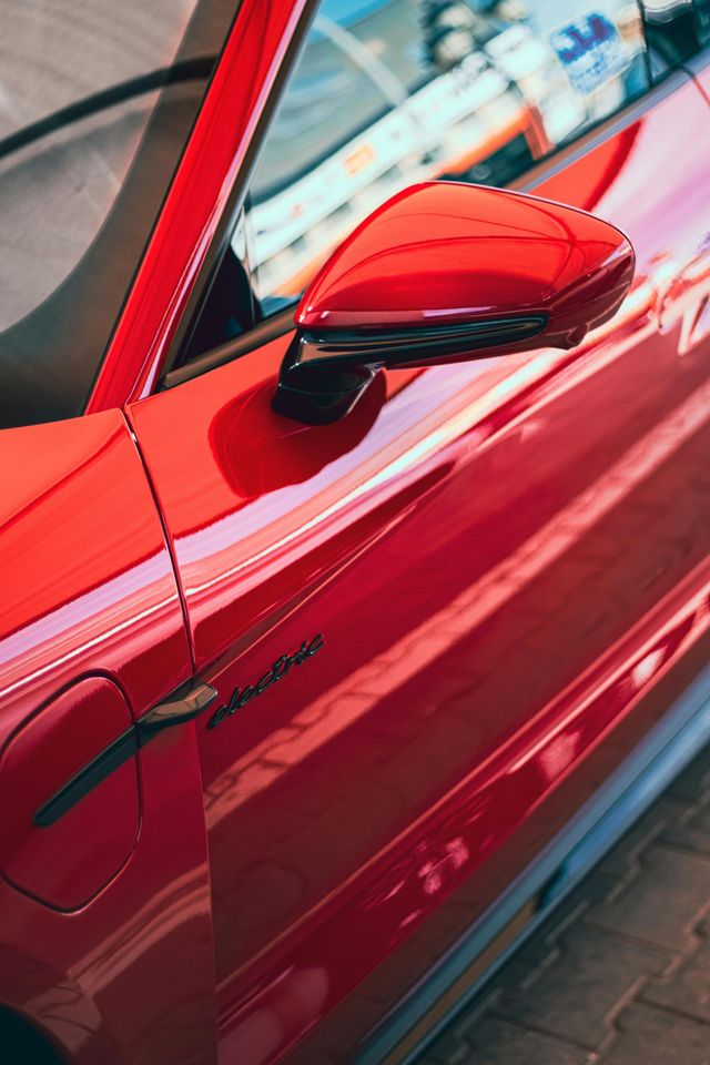 A close-up, high-angle shot of a vibrant red car's side mirror, door panel, and charging port.