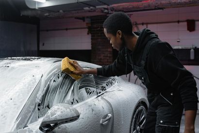 A person in black clothing washes a foam-covered car with a sponge in a garage.